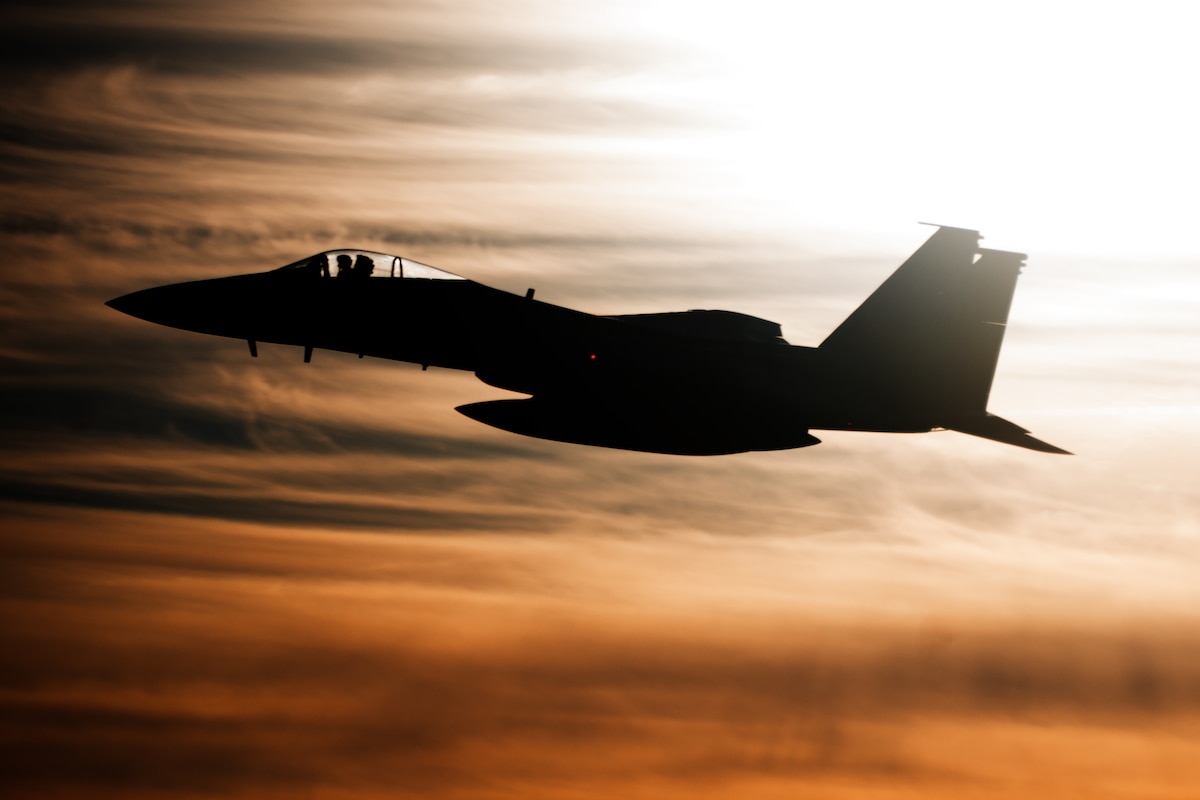 A silhouette of an F-15C Eagle aircraft flies through a burnt orange sky.
