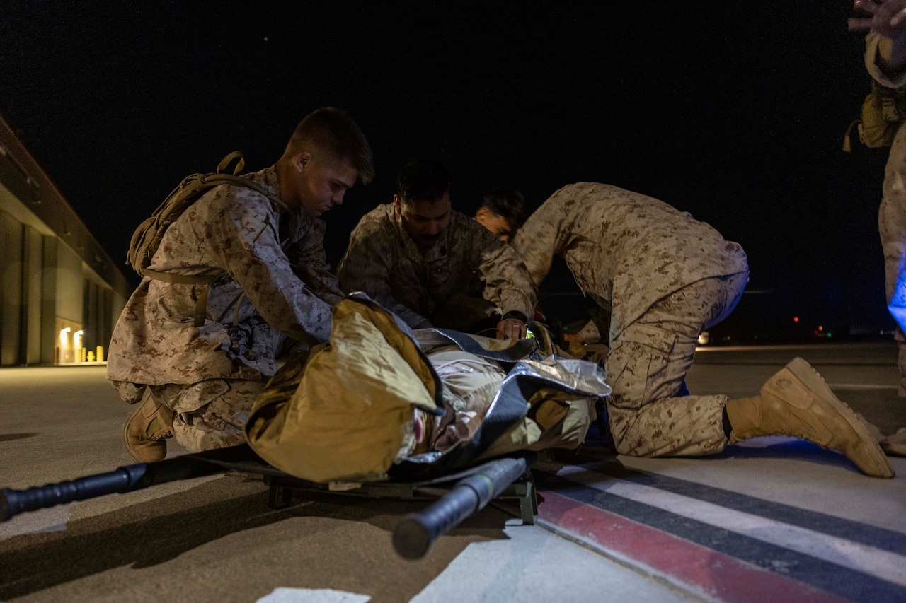 Service members wearing military camouflage uniforms provide medical care to a role player lying on concrete.