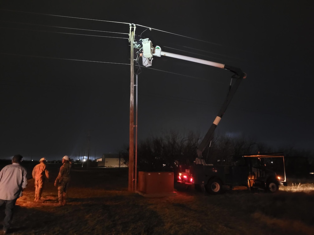 a bucket truck is backed up to a power pole at night. The trucks boom is raised and a light is illuminating the power pole as a worker looks at the power line.