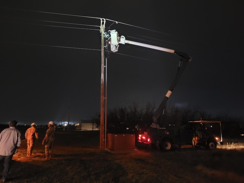 a bucket truck is backed up to a power pole at night. The trucks boom is raised and a light is illuminating the power pole as a worker looks at the power line.