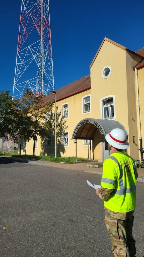 A soldier in a hard hat and reflective vest looks at a communications tower in the distance.