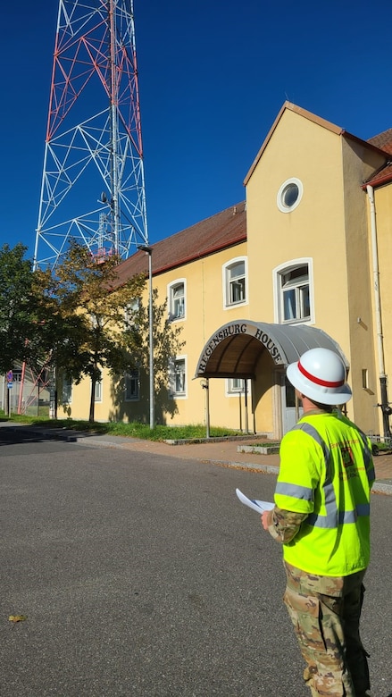 A soldier in a hard hat and reflective vest looks at a communications tower in the distance.