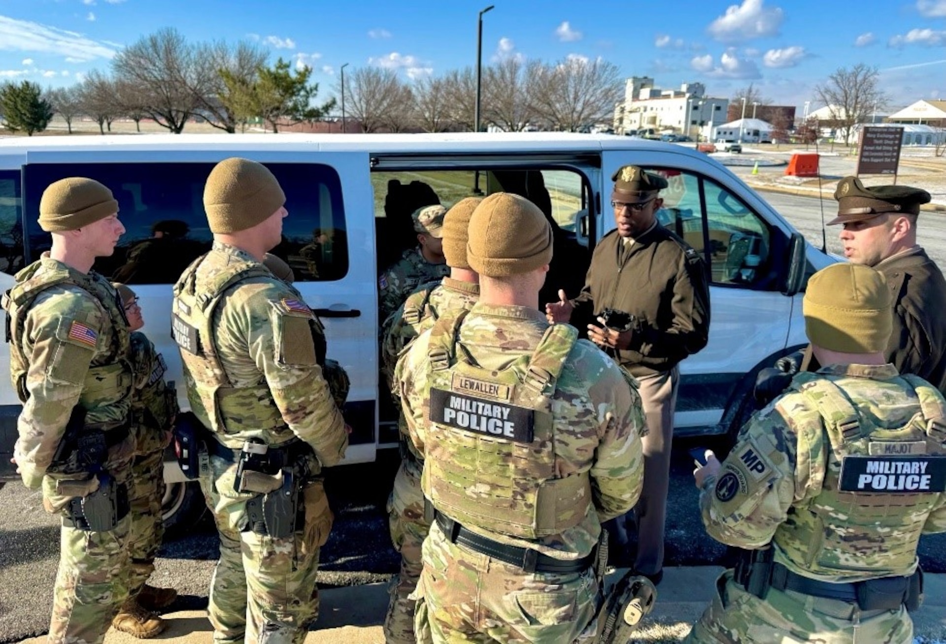 Several Army soldiers wearing combat uniforms with the words Military Police on the back are facing a soldier wearing the Army green service uniform as he talks with them next to a white van.