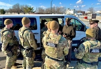 Several Army soldiers wearing combat uniforms with the words Military Police on the back are facing a soldier wearing the Army green service uniform as he talks with them next to a white van.