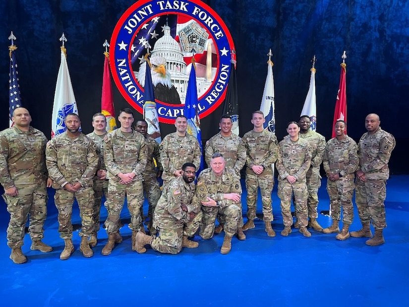 Fourteen Army soldiers in combat uniform are posting for a picture in a room with a blue floor and several flags and a large organizational seal in the background.