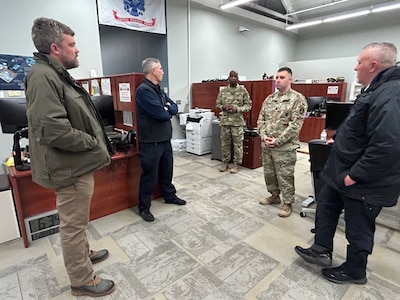 Two Army soldier who are wearing combat uniforms are standing in an office talking with three men in civilian attire.