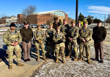 Army Soldiers wearing combat uniforms and Army green service uniforms are standing outside on snow-covered grass posing for a picture with a large arch-roofed building in the background.