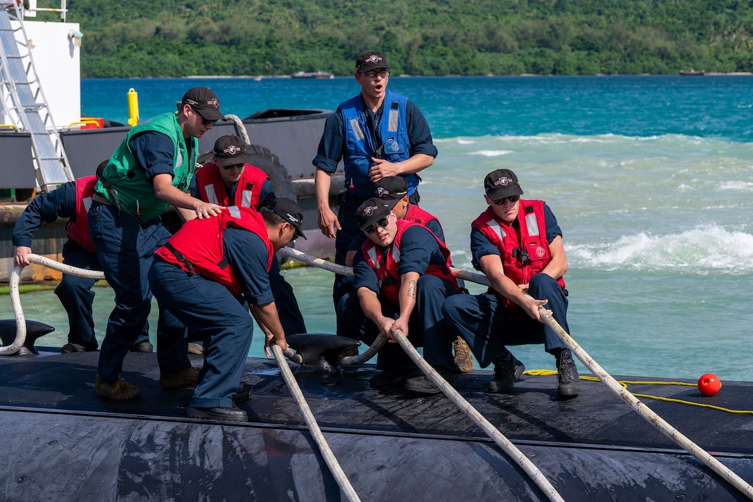 Sailors standing on top of a partially submerged submarine pull on large cables during daytime, with a body of water and land with green vegetation in the background.