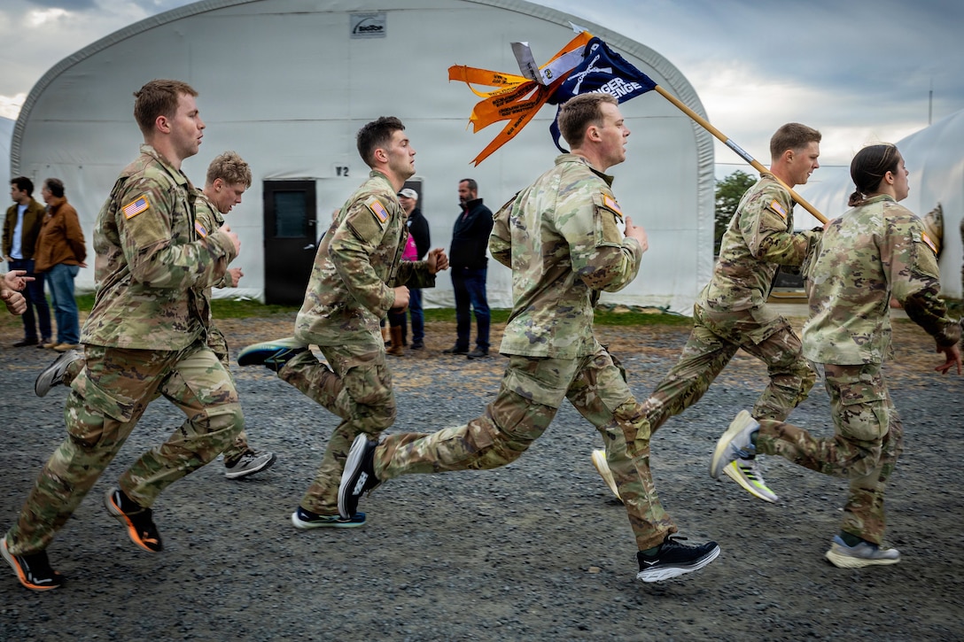 ROTC cadets in camouflage uniforms, one carrying a pole with a flag, run on a rocky path under a cloudy sky, with spectators and large canopy structures in the background.