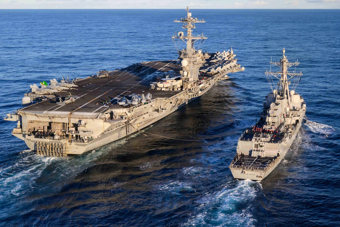 An aircraft carrier and a smaller ship are connected by a large cable while sailing in a large body of water during daytime.
