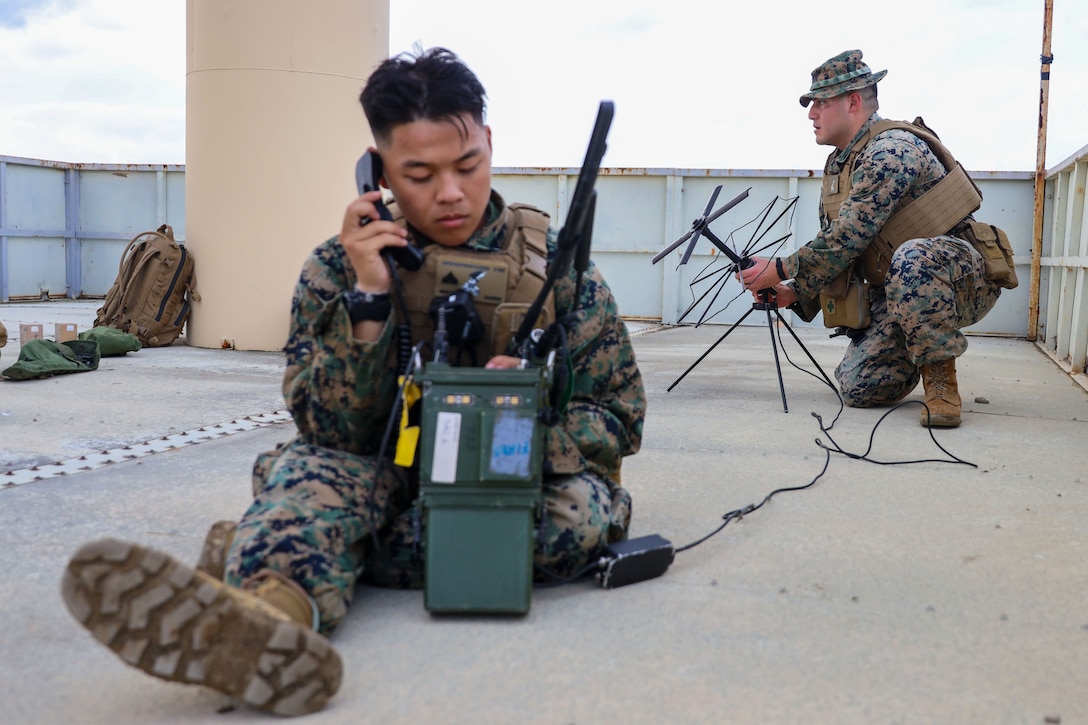 Two Marines, one sitting and holding a phone device to an ear and adjusting an attached piece of equipment and the other crouching while adjusting an antenna, are seen inside an open structure under a cloudy, daytime sky.