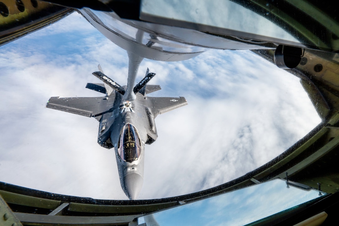A flying jet approaches a piece of equipment attached to another aircraft above it, with clouds in the background.