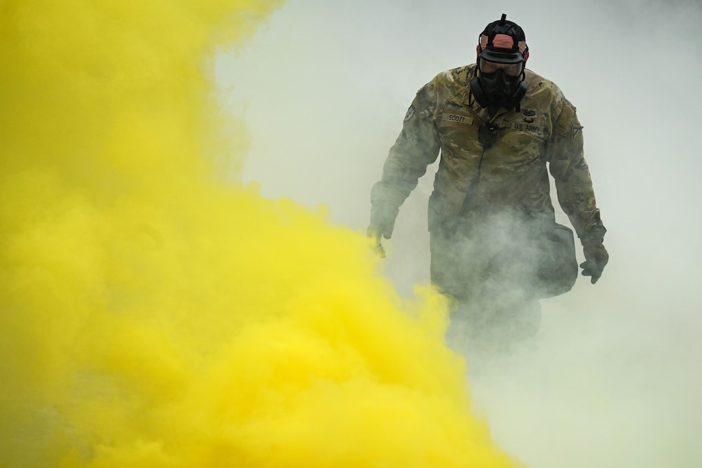A soldier wearing a gas mask and camouflage uniform looks downward as yellow and white smoke swirls.