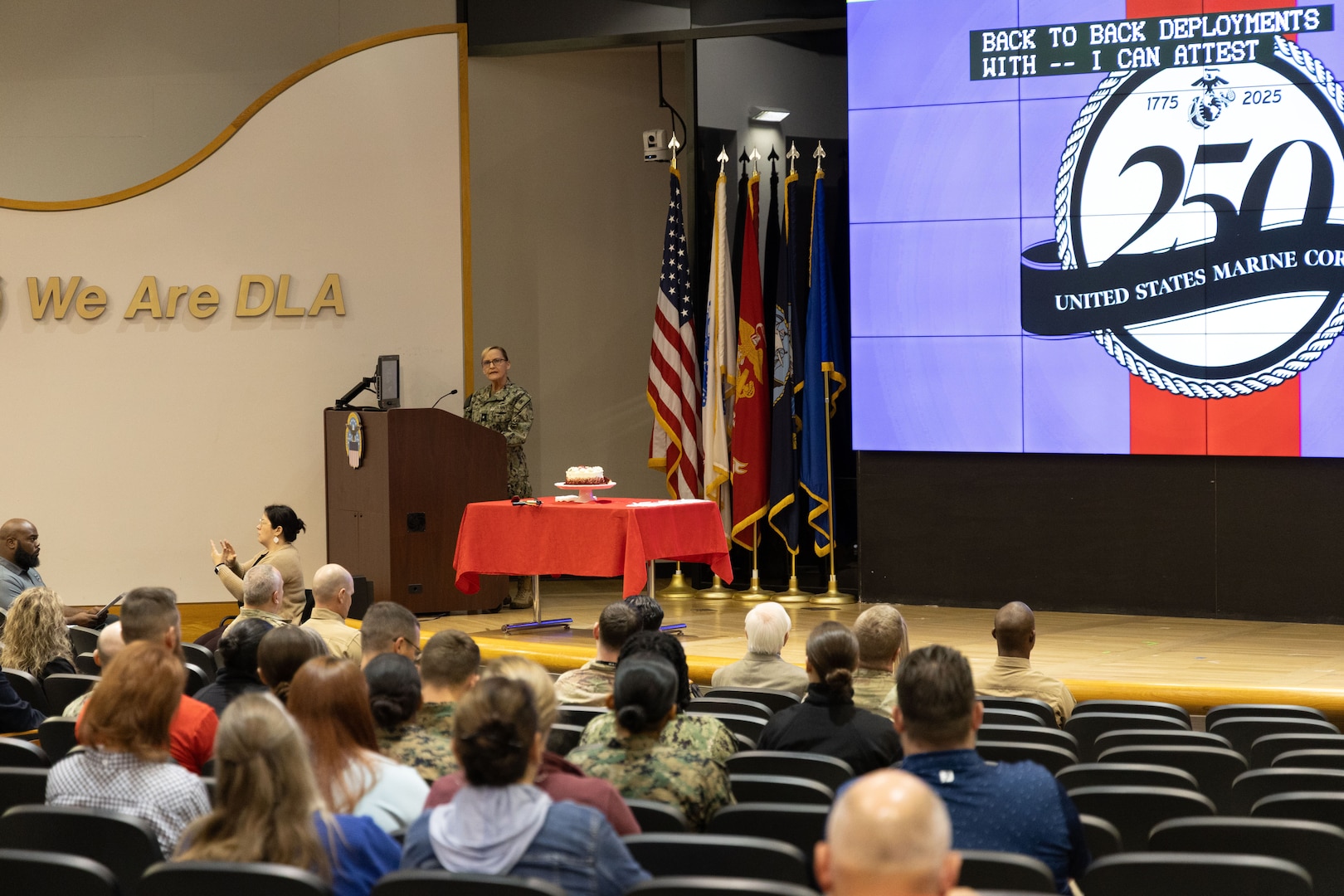 Woman on stage behind podium to the left speaks to audience