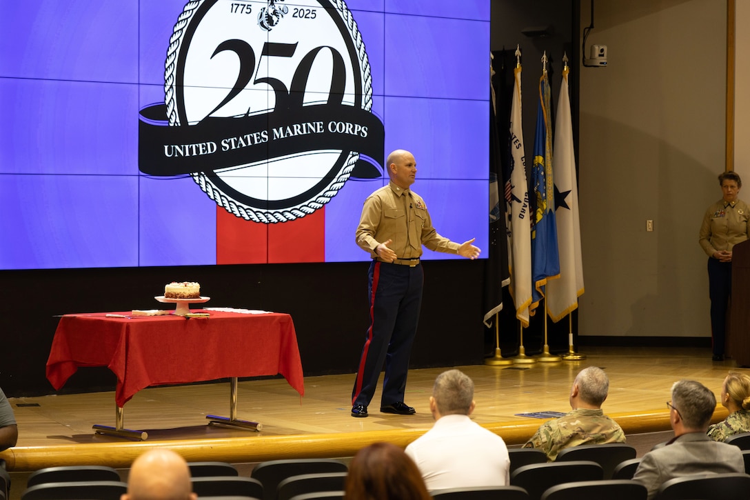 Marine stands on stage speaking to audience - cake on table to the left