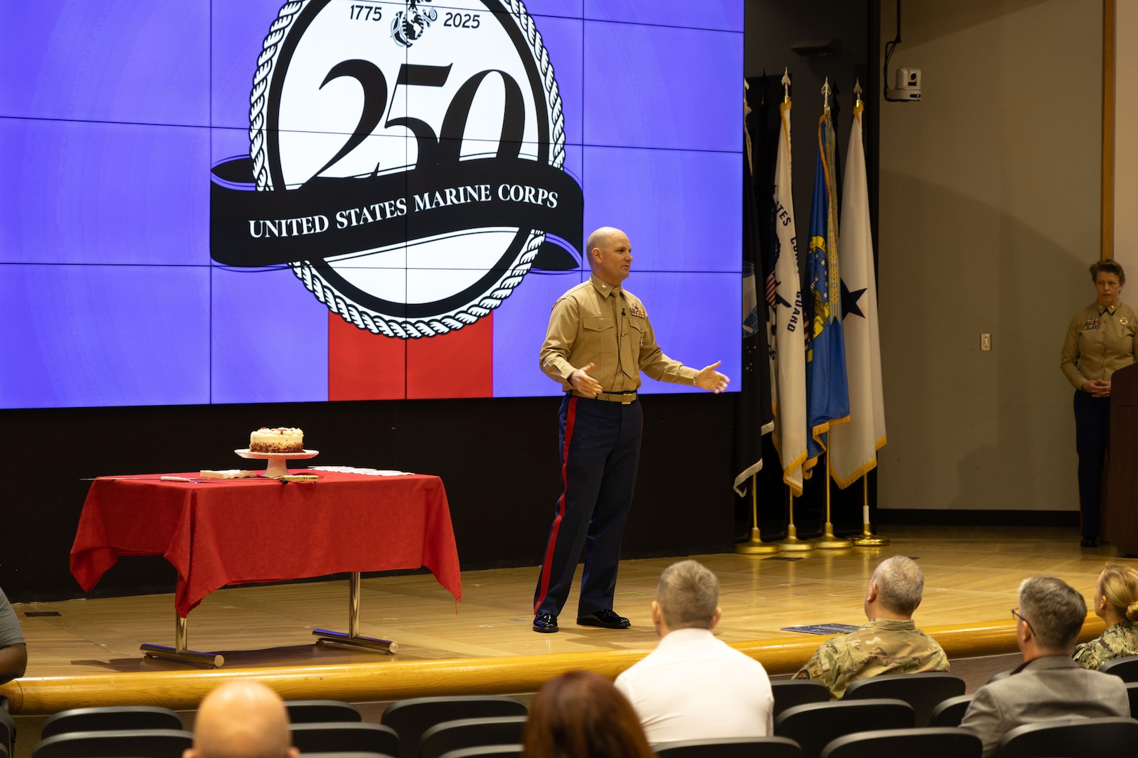 Marine stands on stage speaking to audience - cake on table to the left