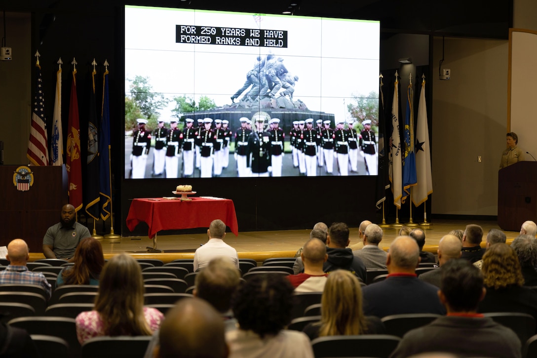 People seated in auditorium watch large screen above stage with Marines on display