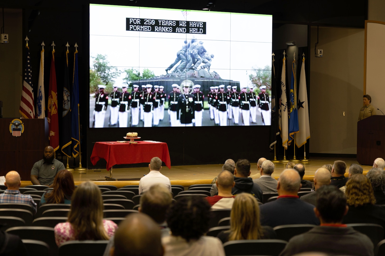 People seated in auditorium watch large screen above stage with Marines on display