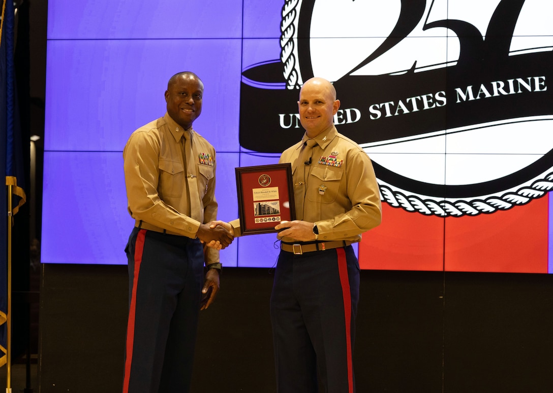Two men in Marine uniform shake hands and pose for photo. Man on right holing a frame