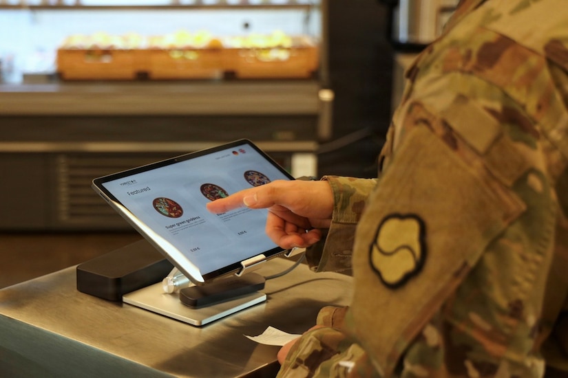 A soldier wearing a camouflage military uniform chooses a meal on a touchscreen tablet.