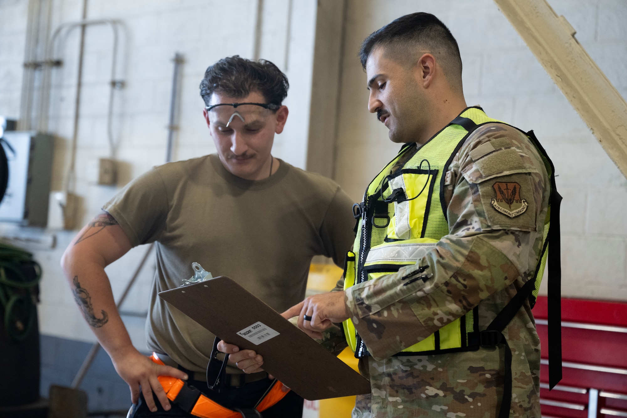 A man in a military uniform and a bright yellow vest points at a clipboard next to another man wearing mechanic type clothing