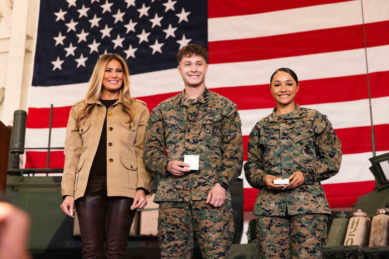 A woman wearing business casual attire stands next to two Marines in military camouflage uniforms holding small boxes in their hands. A large American flag is in the background.