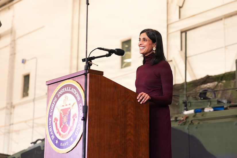 A woman wearing business attire stands in front of a lectern while speaking into a microphone.