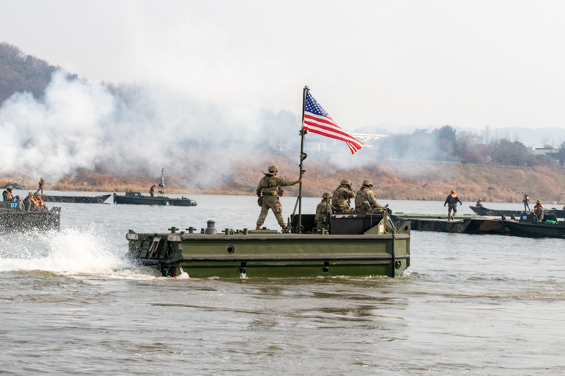 Soldiers aboard a boat flying an American flag transit a body of water as other boats converge in the area, with lingering smoke and tree-scaped land in the background.