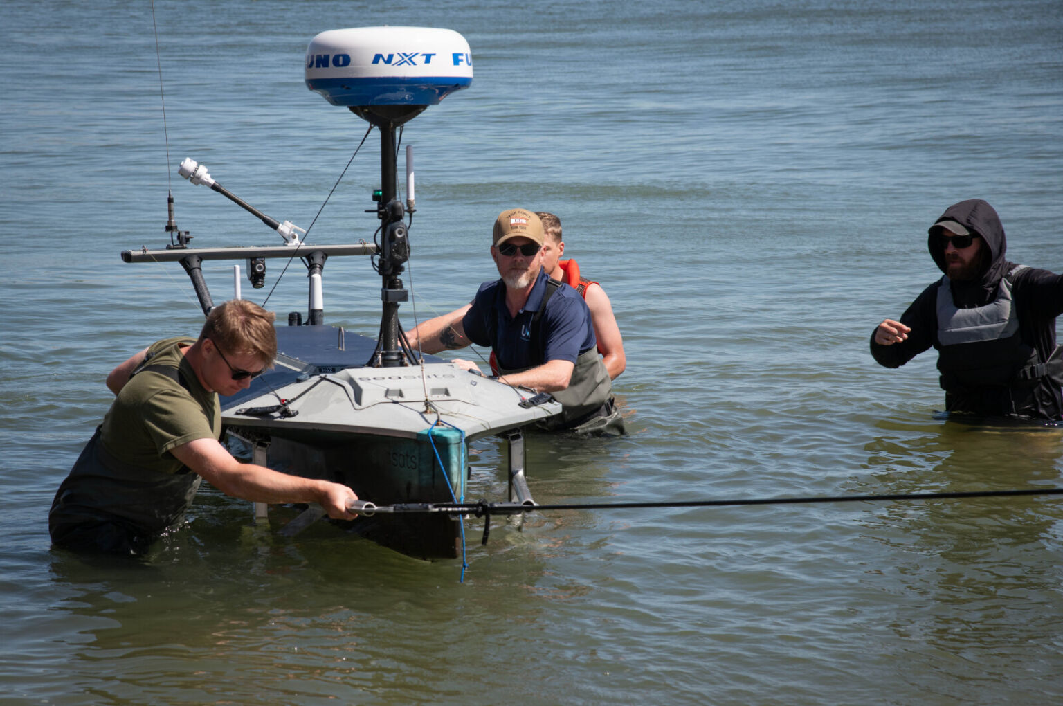 Three men in waterproof coveralls guide an unmanned surface vessel through waist-deep water. Another man in similar attire observes them.