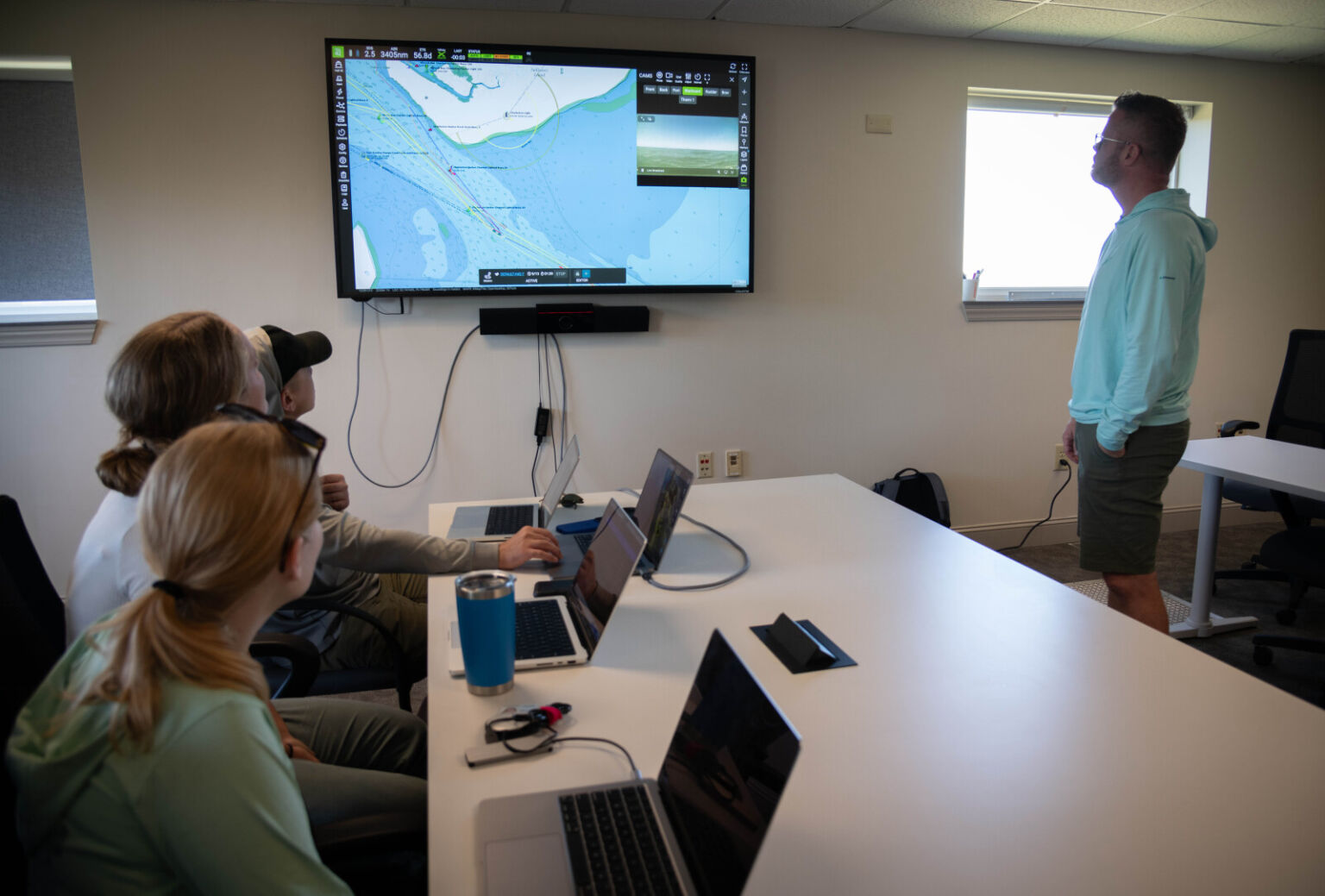 Three people seated at a table with laptops in front of them and a man standing on the other side of the table look at a screen on the wall that is displaying a map.