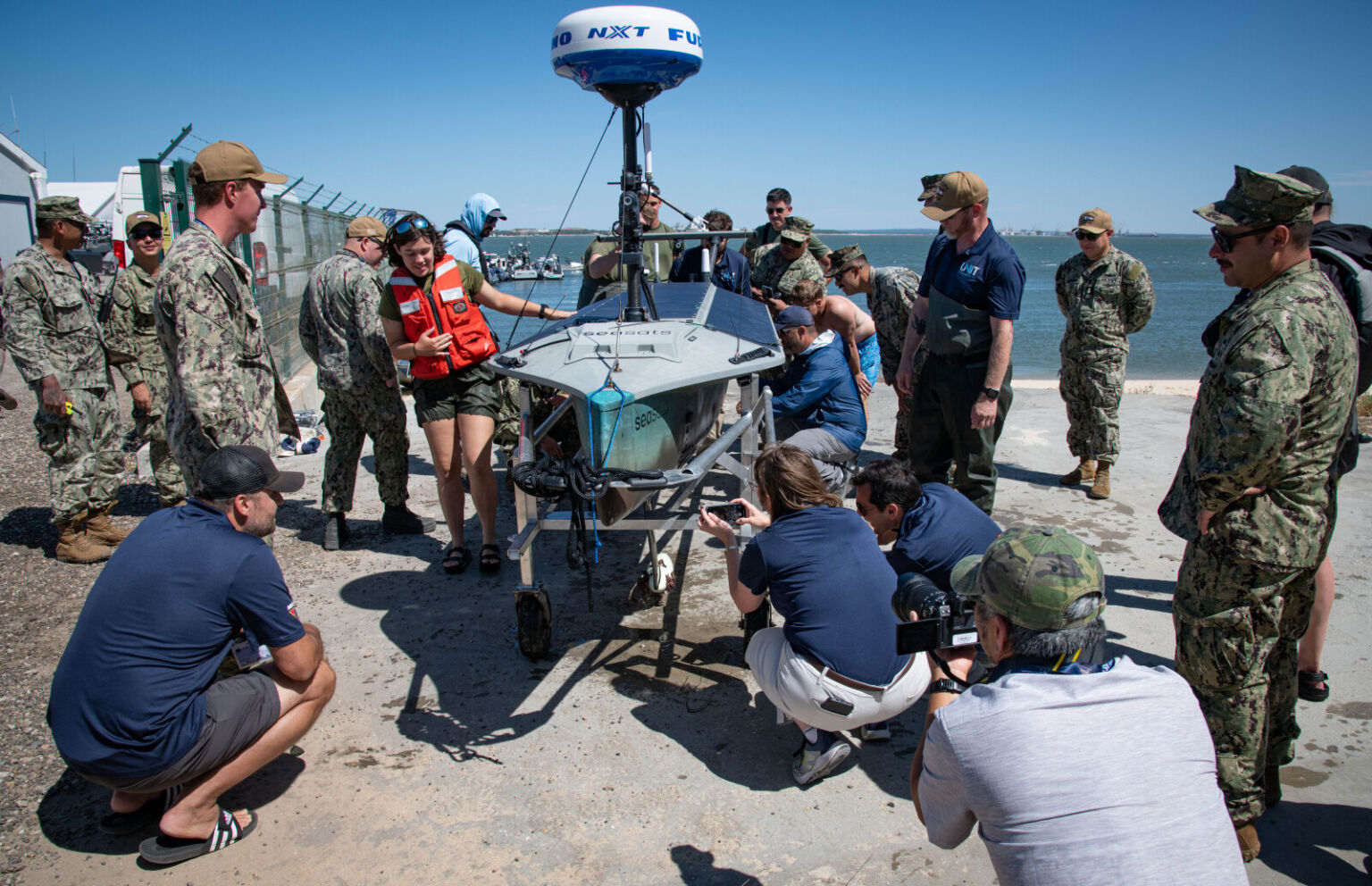 Several people in business casual attire and military camouflage uniforms gather around an unmanned surface vessel as they prepare to launch it into the water.