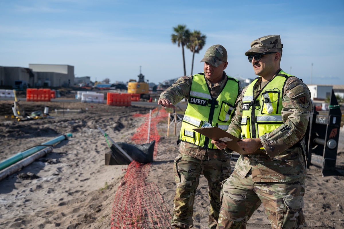 Two men wearing military uniforms and yellow vests look at a construction site. One is pointing while the other holds a clipboard
