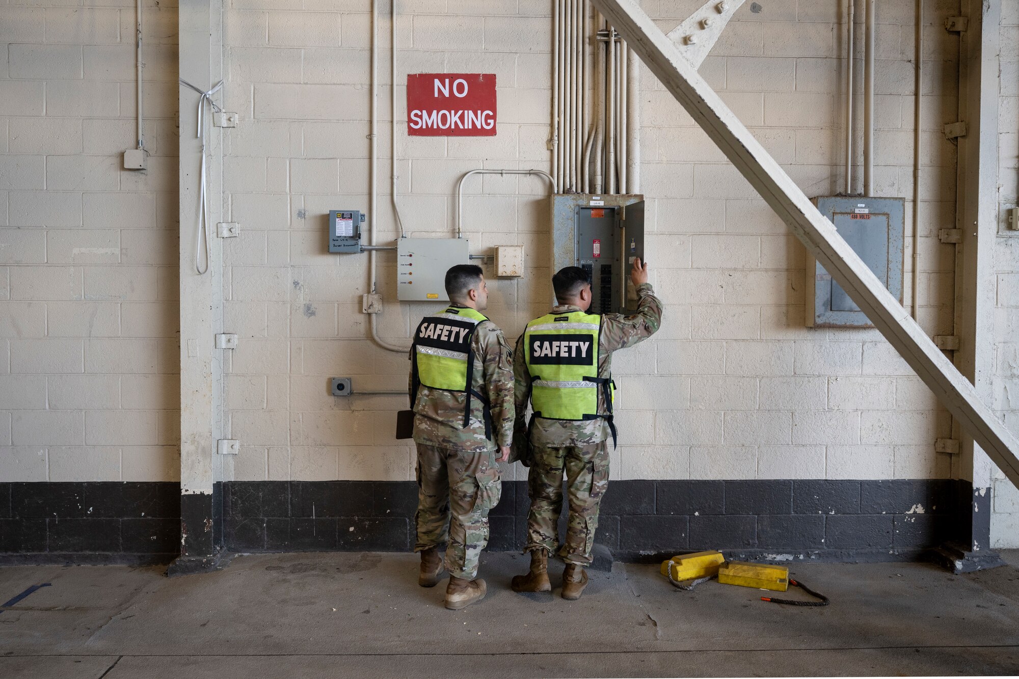 Two men wearing yellow vests and military clothing stand in the center of the photo looking at an electrical box