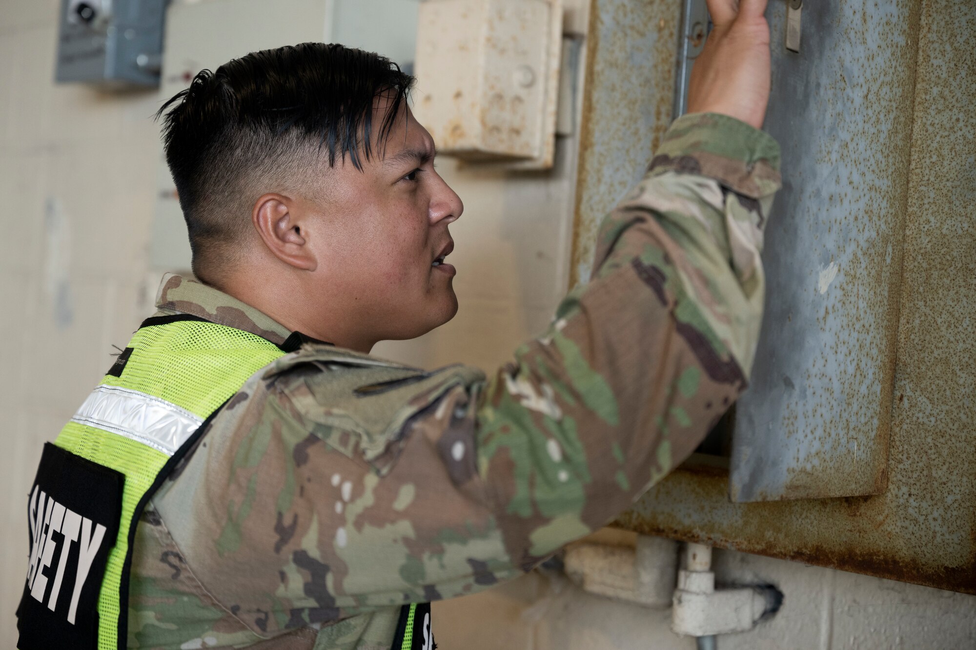 A man wearing a yellow vest over military clothing looks at an electrical box