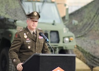 A soldier standing at a podium speaking with an Army motor vehicle in the background with camouflaged netting.