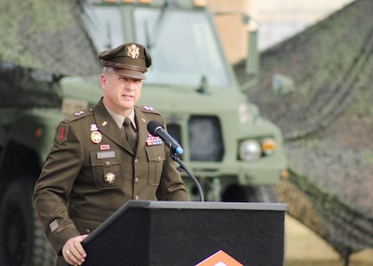 A soldier standing at a podium speaking with an Army motor vehicle in the background with camouflaged netting.