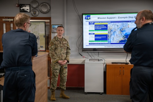 Three military members discuss a slide shown on a television screen.