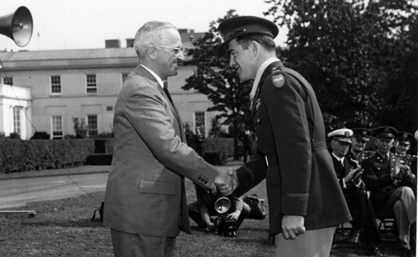 Two men smile and shake hands on a lawn. Others sit in chairs in the background, clapping.