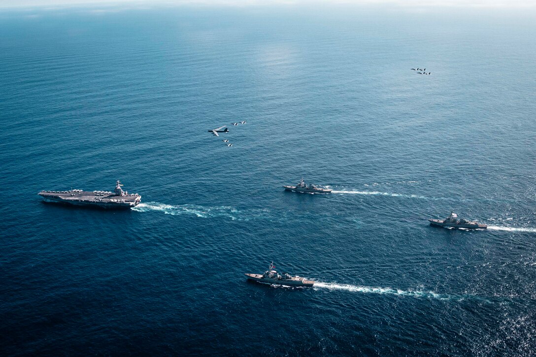 An overhead view of a group of four Navy warships steaming across the ocean in the daytime. A handful of military aircraft fly above them.