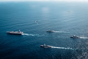 An overhead view of a group of four Navy warships steaming across the ocean in the daytime. A handful of military aircraft fly above them.