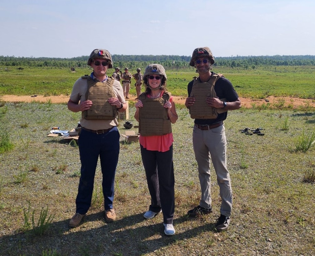Three individuals in Marine Corps helmets and Kevlar pose for a photo in a field.