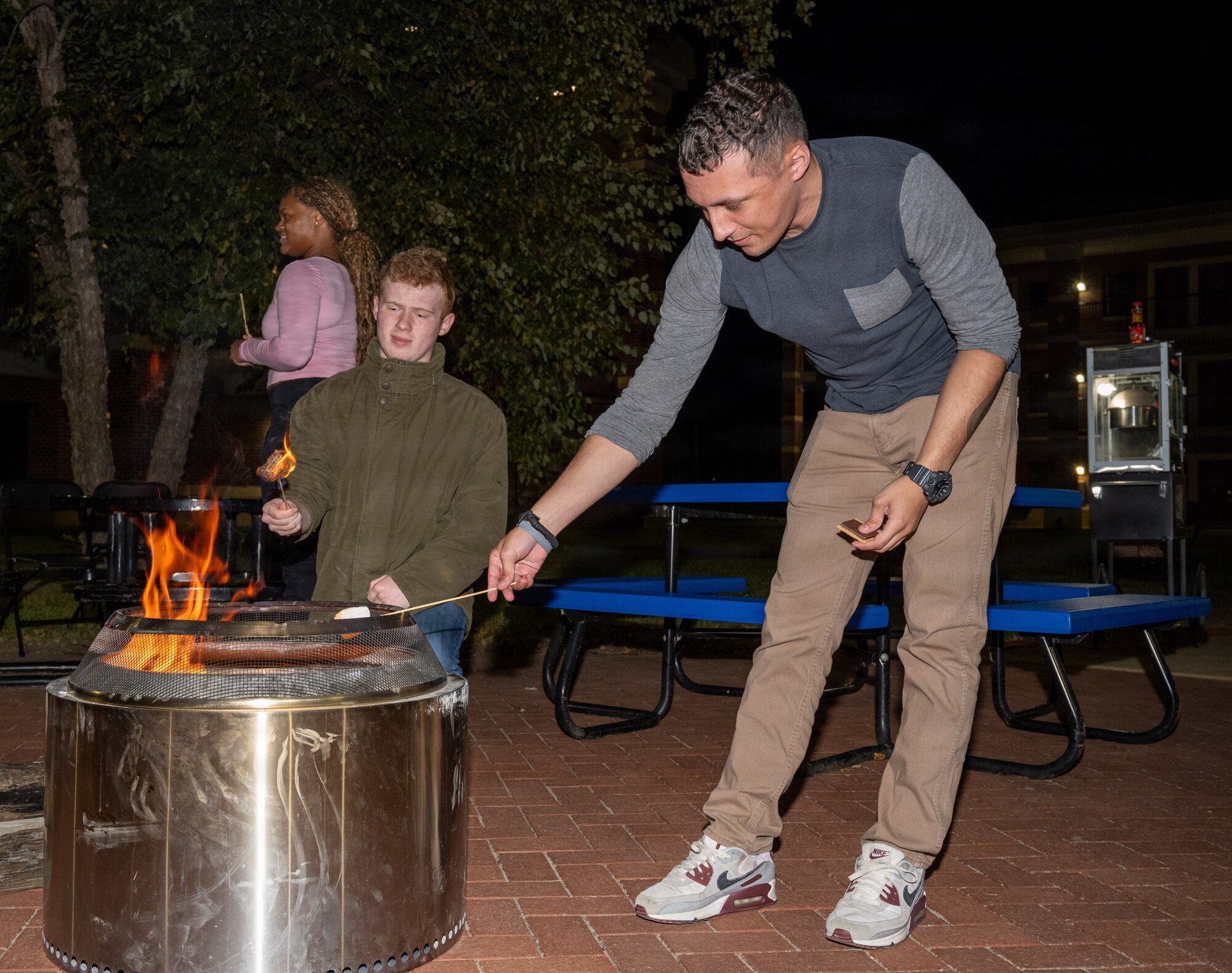 Two men hold marshmallows over a metal pit with fire.