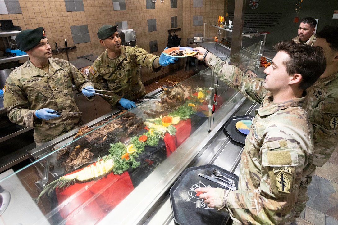 Two soldiers wearing green berets and camouflage uniforms serve food on a plate to three other soldiers with trays and silverware in a cafeteria setting.