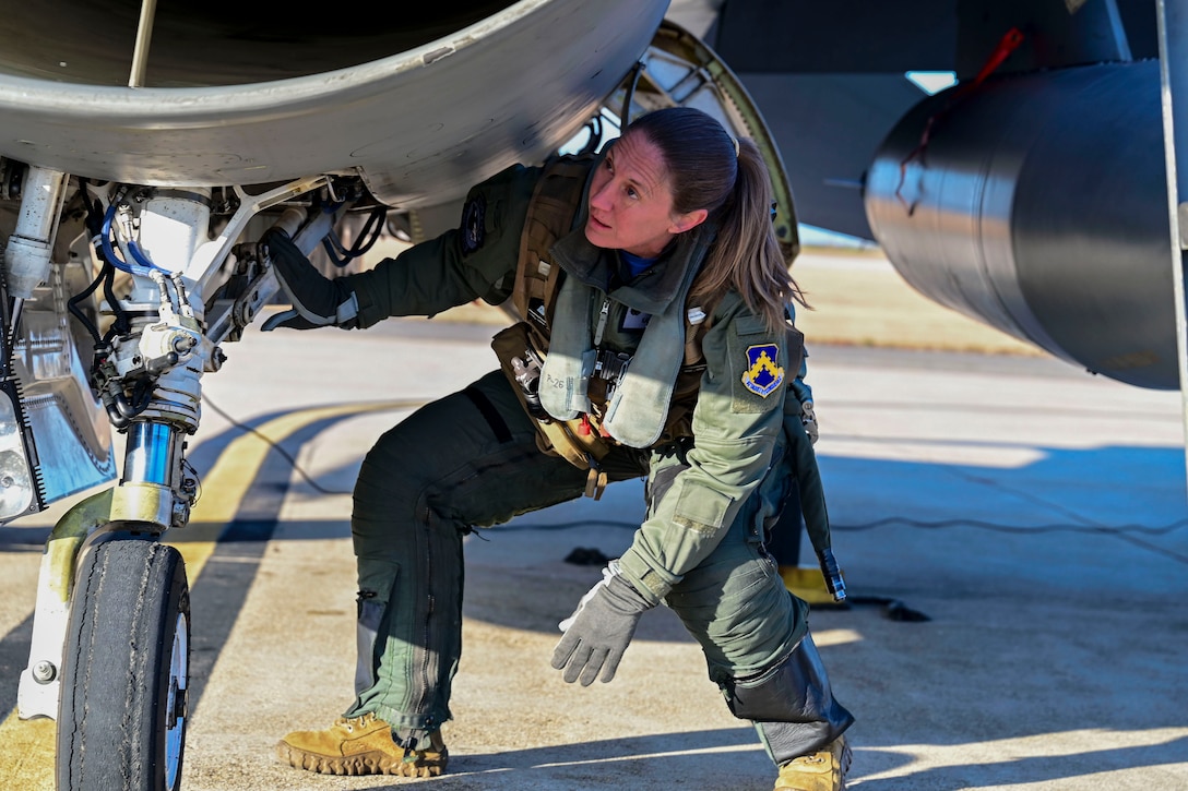 An airman wearing a green flight suit crouches to inspect the underside of an aircraft on a runway during daytime.