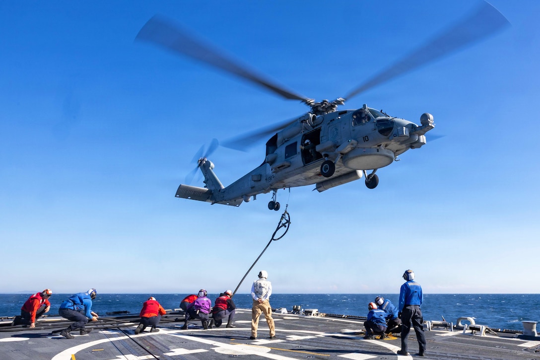 Sailors crouch and stand on the deck of a ship as a helicopter hovers above, with a person in the open door of the helicopter guiding a cable up from the deck on a cloudless day; a body of water is in the background.