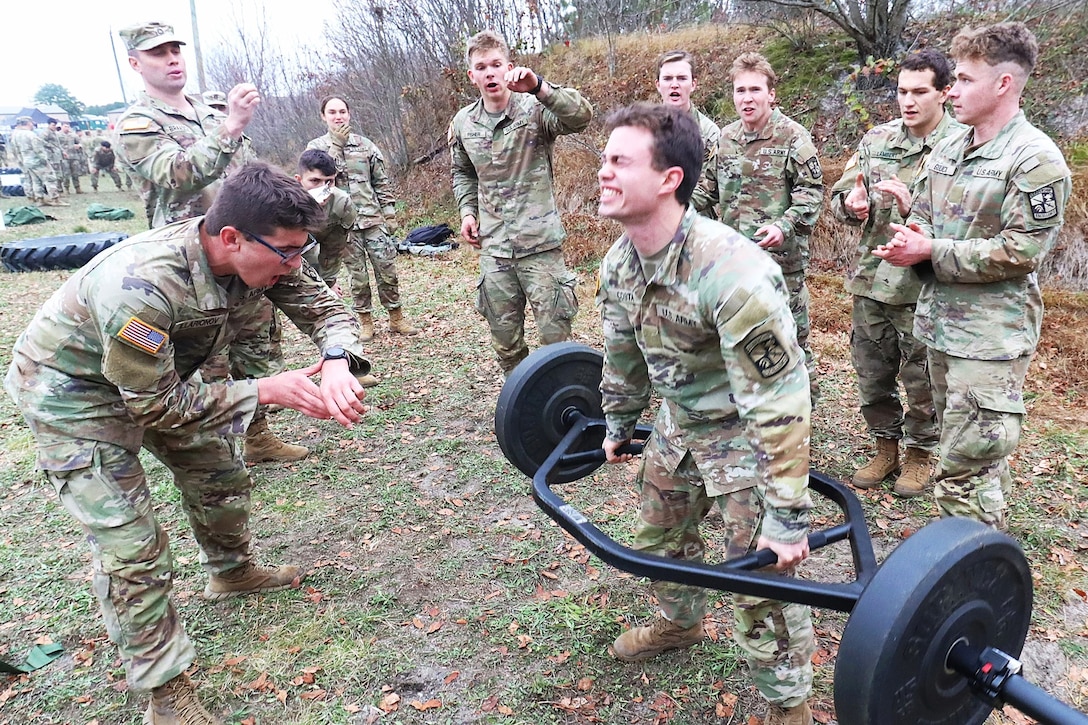 Army ROTC cadets cheer on another cadet performing a dead lift with a weight set on grass during daytime, with another group of cadets and trees in the background.
