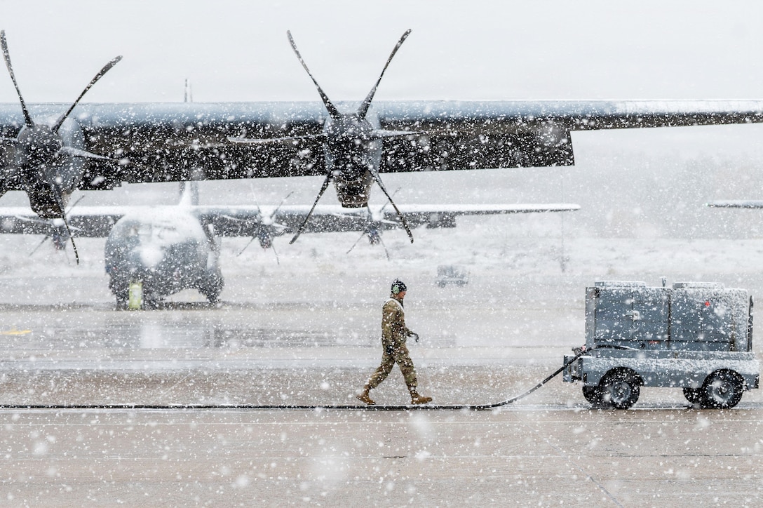 An airman walks toward a piece of equipment with four tires and a long cable attached to it on a runway on a snowy day, with two large planes in the background.