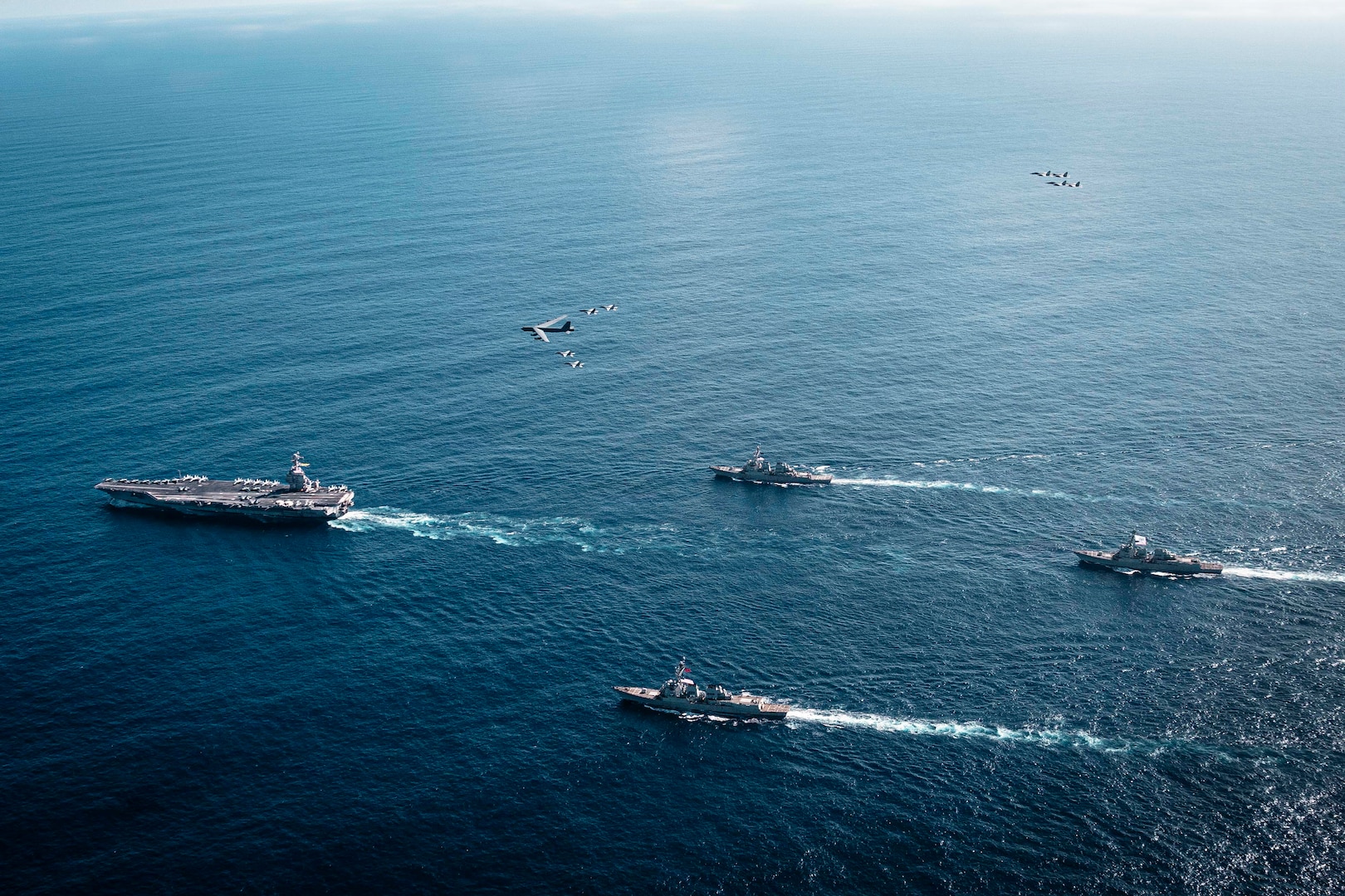An overhead view of a group of four Navy warships steaming across the ocean in the daytime. A handful of military aircraft fly above them.