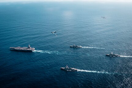 An overhead view of a group of four Navy warships steaming across the ocean in the daytime. A handful of military aircraft fly above them.