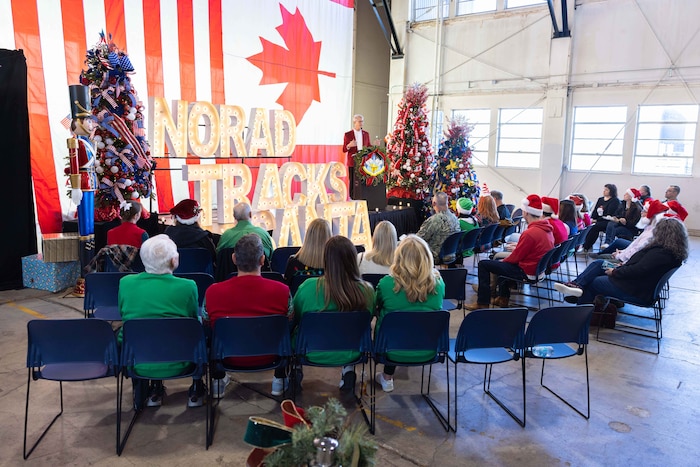 A person in business attire speaks behind a lectern to a group of seated people in holiday attire. There are holiday decorations displayed around the stage.